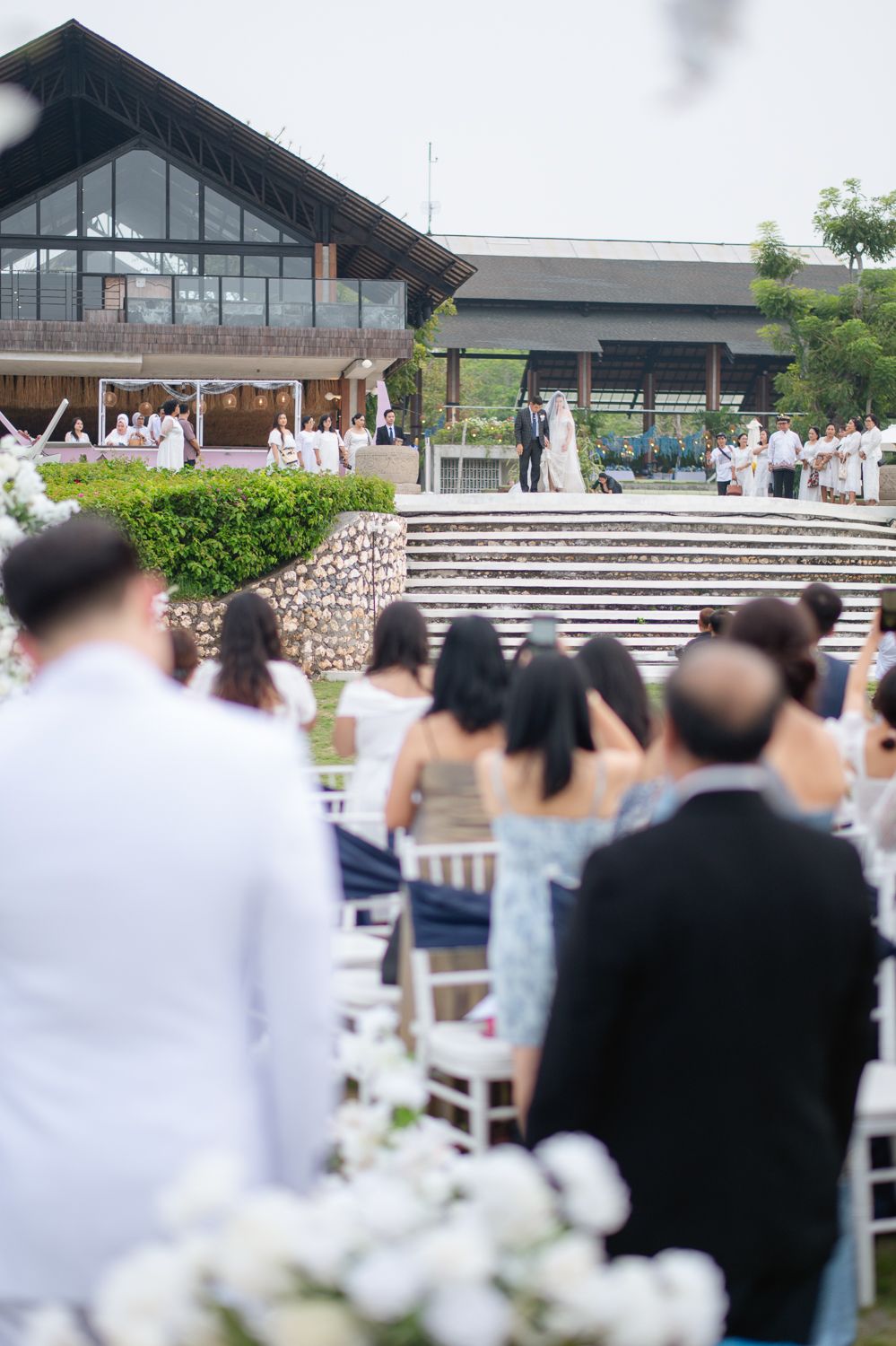 Outdoor wedding ceremony with guests seated on white chairs facing couple at altar steps before elegant venue.