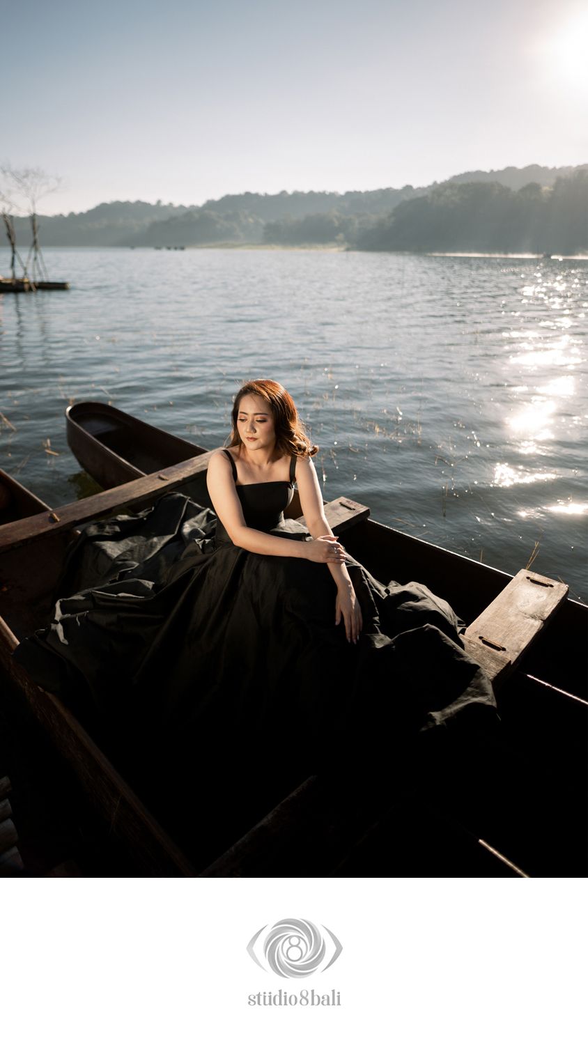 Woman in black gown sitting gracefully in a wooden boat on a sunlit lake.