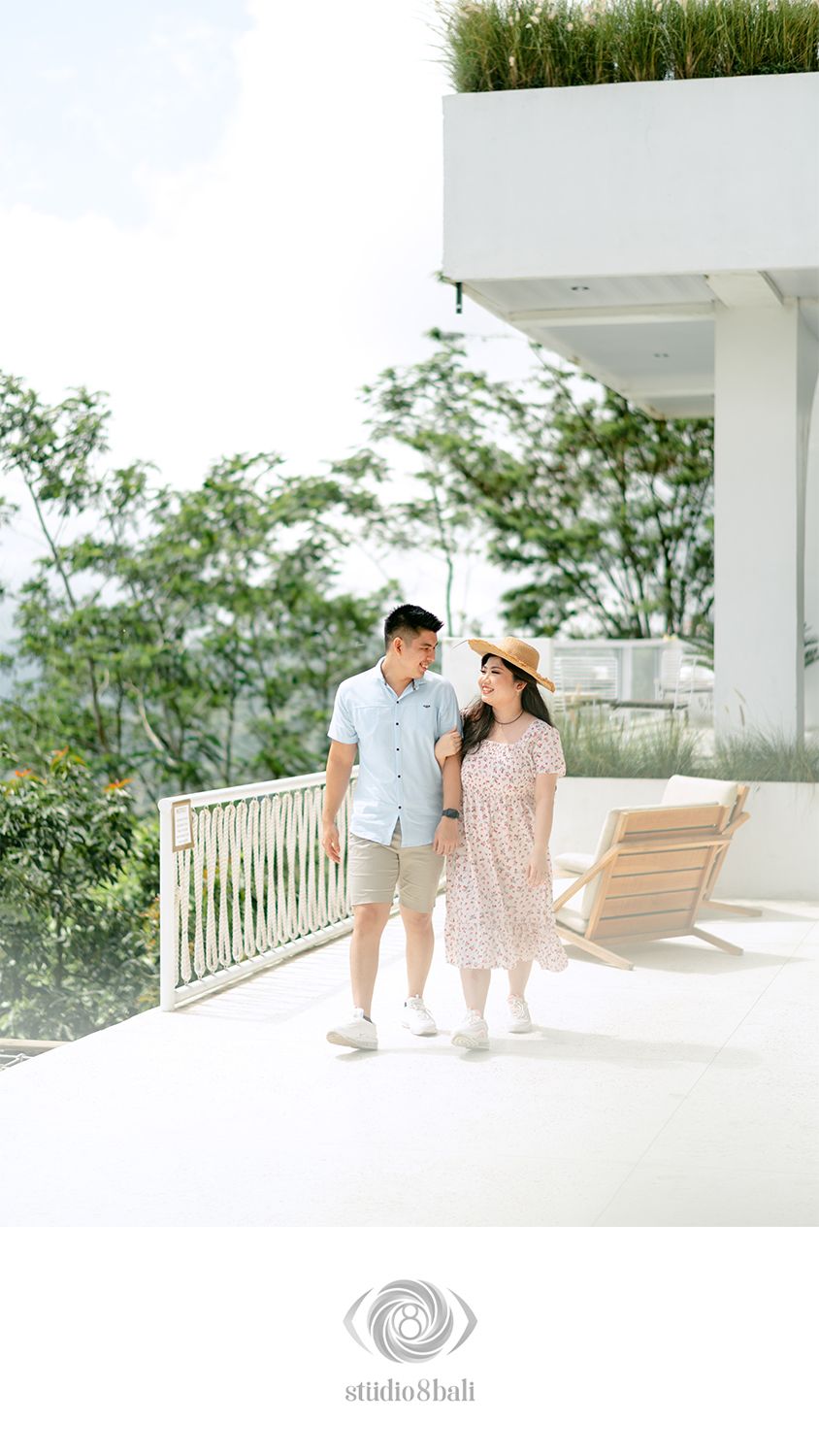 A couple walks hand-in-hand on a white terrace, surrounded by lush greenery, the woman wearing a floral dress and straw hat.