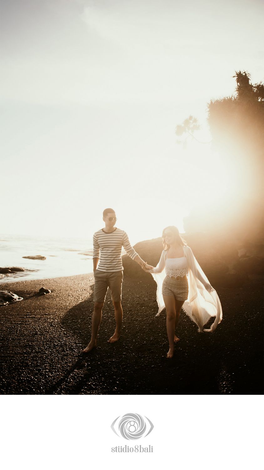 Couple holding hands walking on a black sand beach at golden sunset, captured by Studio 8 Bali.