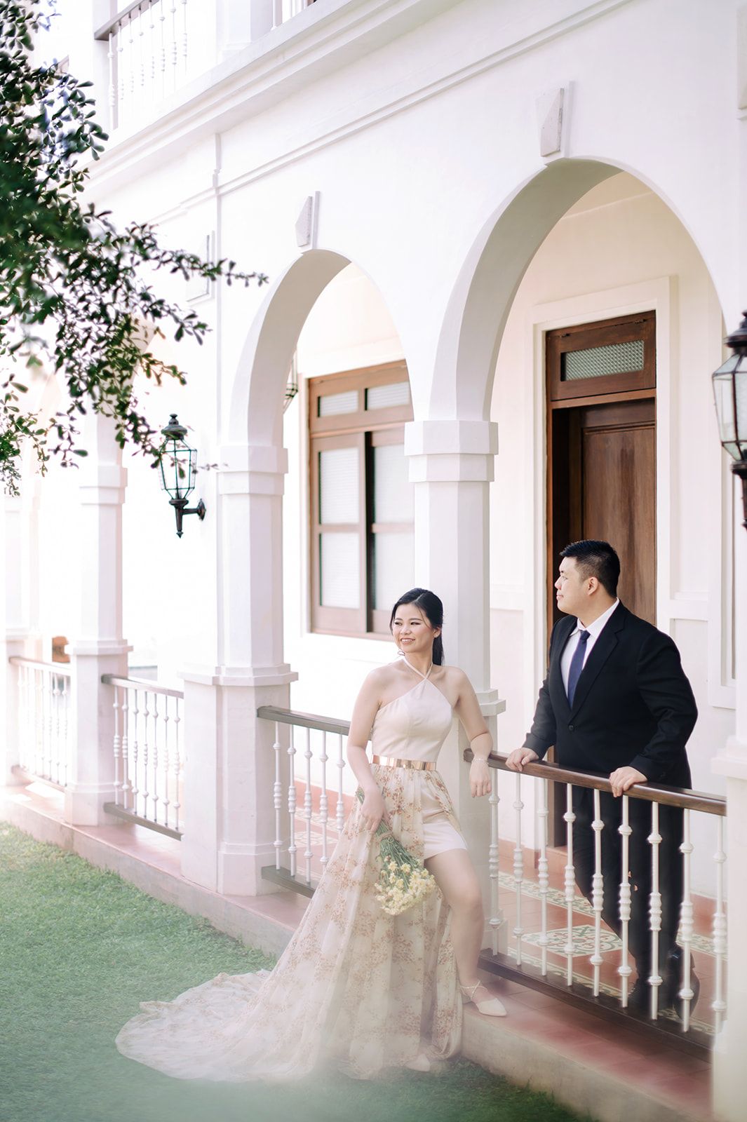 Bride in lace gown and groom in black suit on white colonial building balcony.