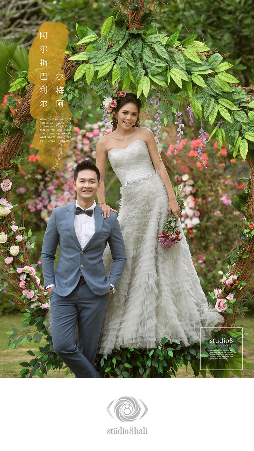 Couple in wedding attire posing before a floral arch with greenery, man in blue suit, woman in white strapless gown holding bouquet.