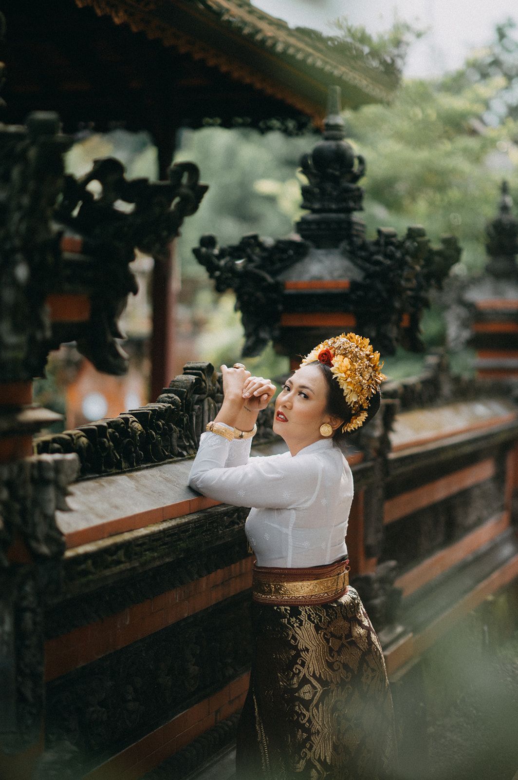 A woman in traditional Balinese attire with a floral headdress poses gracefully at an ornate temple railing.