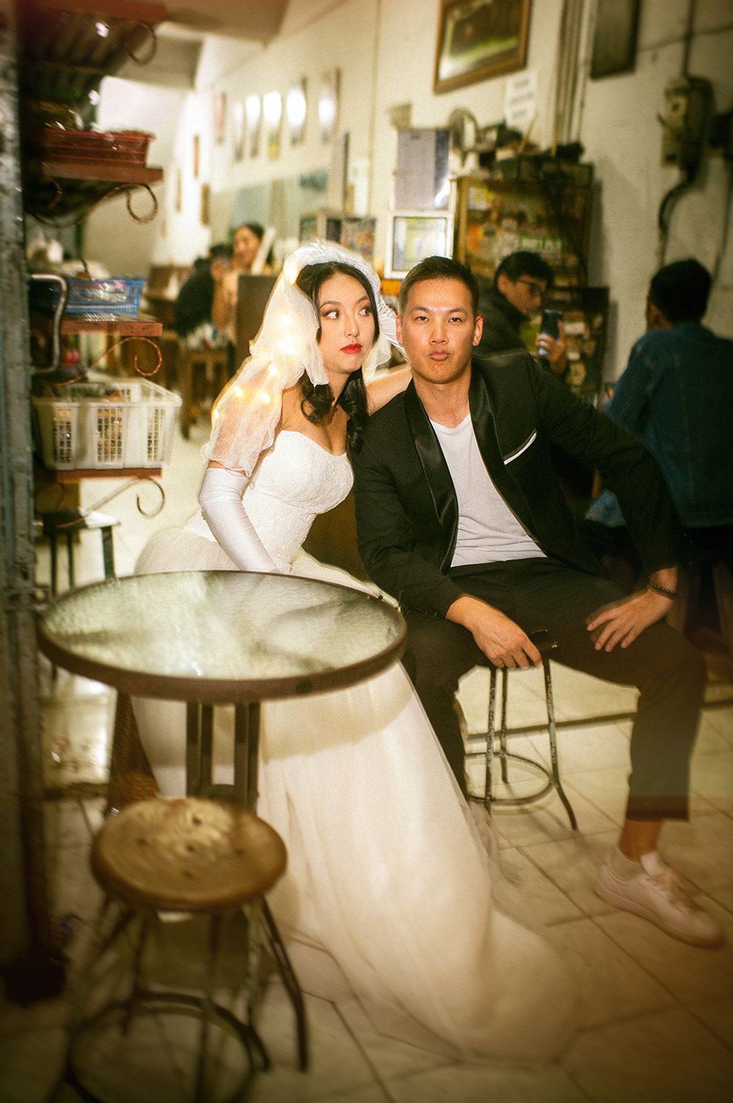 A couple in wedding attire poses in a vintage cafe, the bride in white gown and groom in black suit making playful faces.