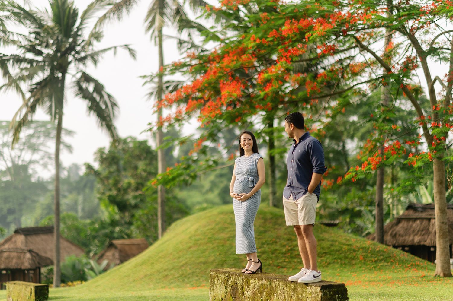 Pregnant couple standing on stone ruins surrounded by lush tropical greenery and vibrant orange flowering trees in Bali Maya Ubud Resort an.