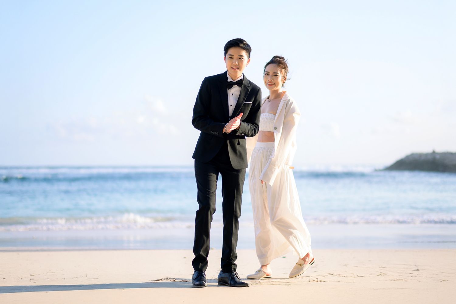 Couple in wedding attire posing on a sandy beach with blue ocean waves and clear sky in the background.