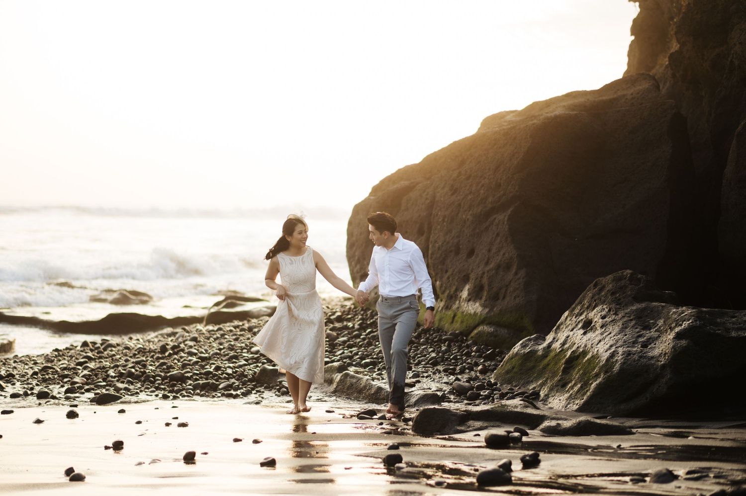A couple holds hands and walks along a rocky beach at sunset, with waves and golden light in the background.