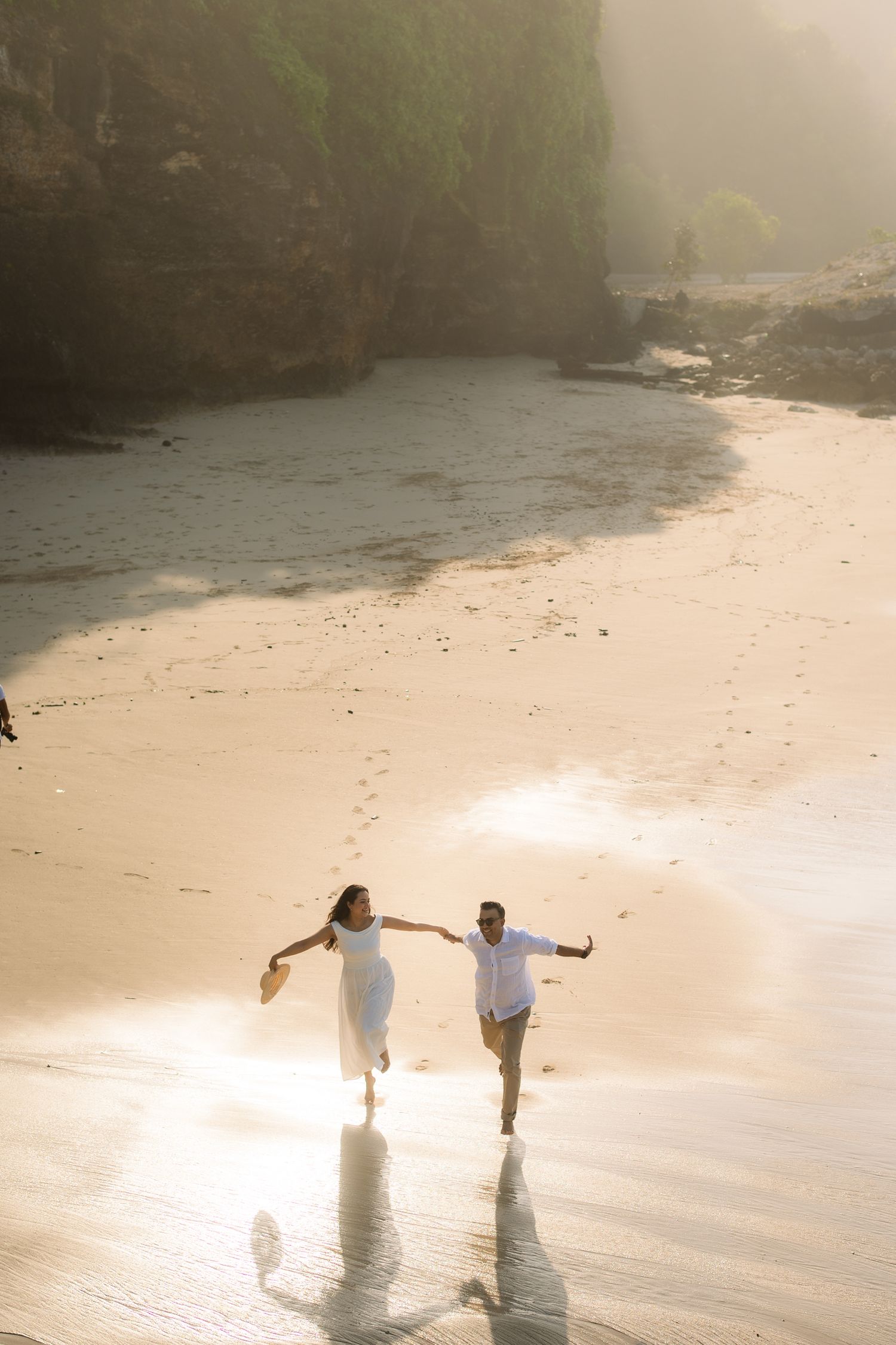 Couple in white running joyfully on a sunlit sandy beach with dramatic cliffs in the background.