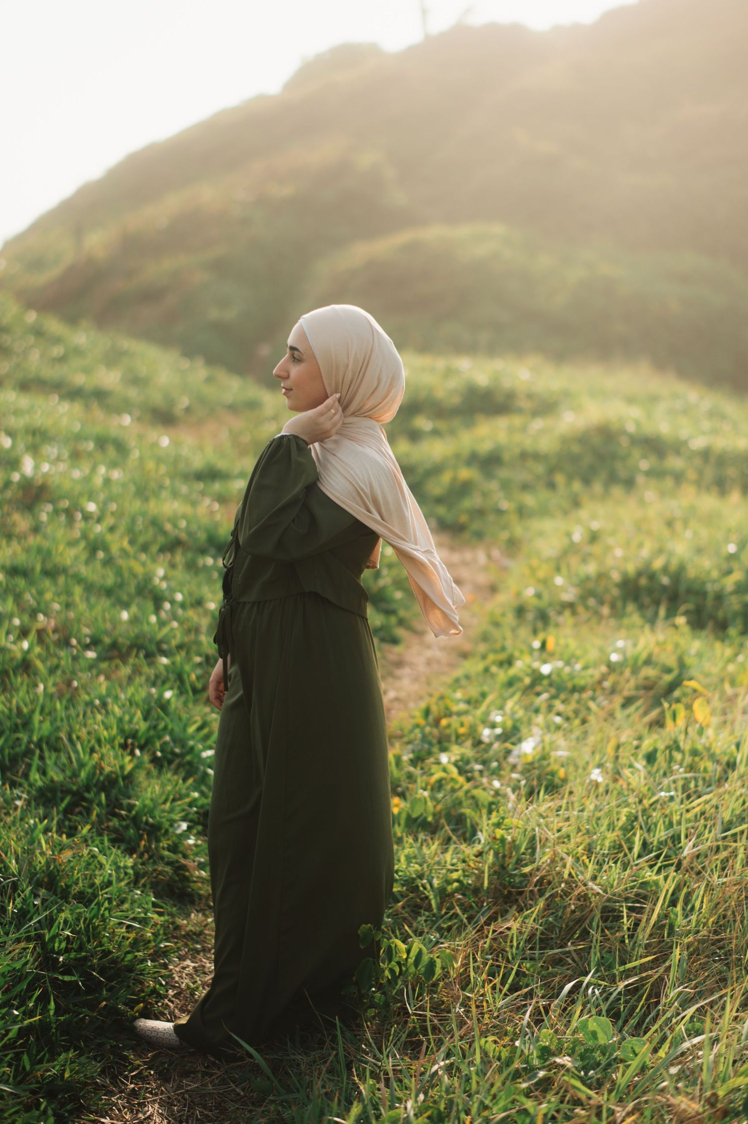 Woman in dark green abaya and beige hijab standing on lush green hillside in golden sunlight.