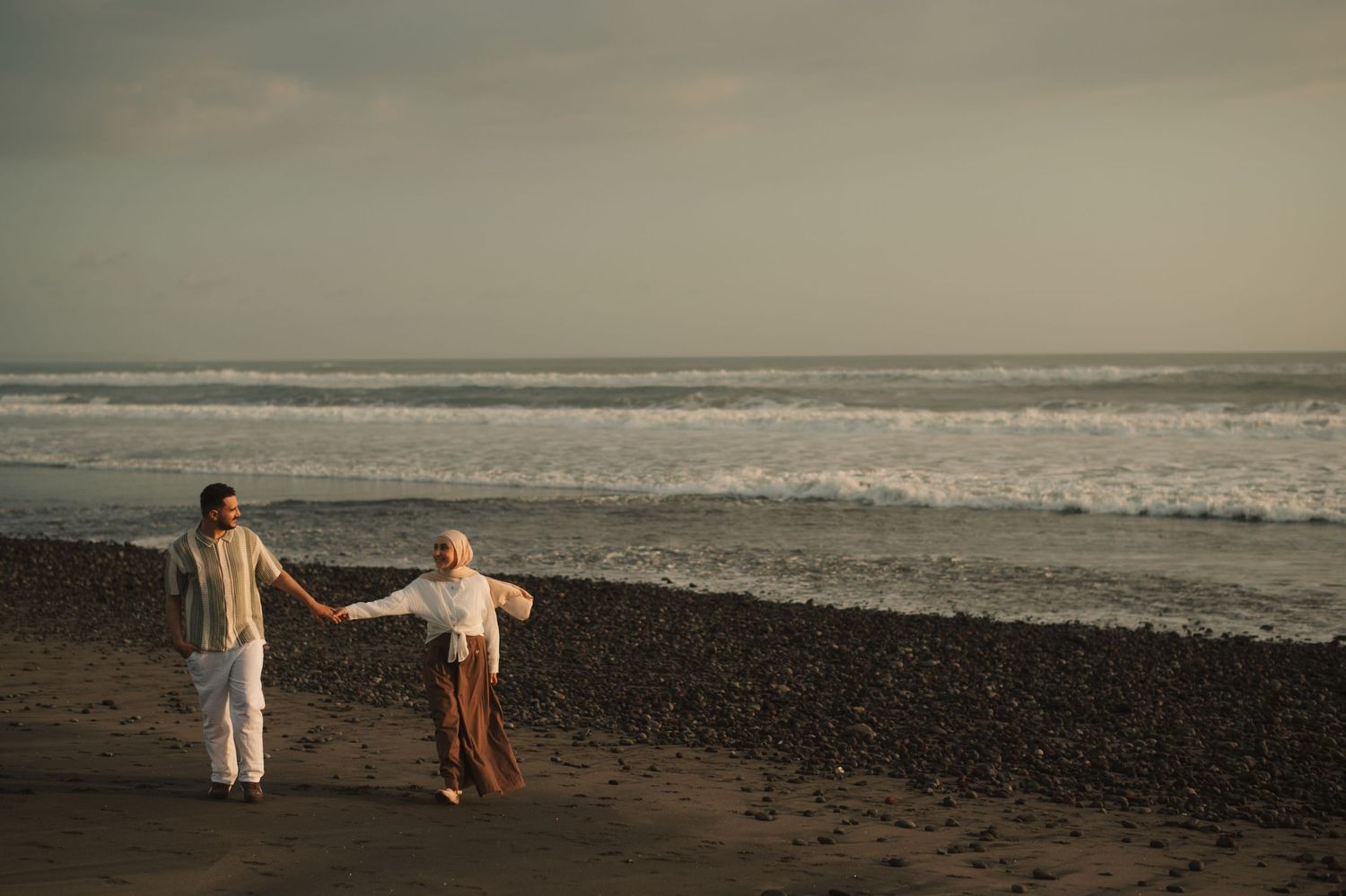 Couple holding hands walking along a dark sandy beach with ocean waves in the background at dusk.