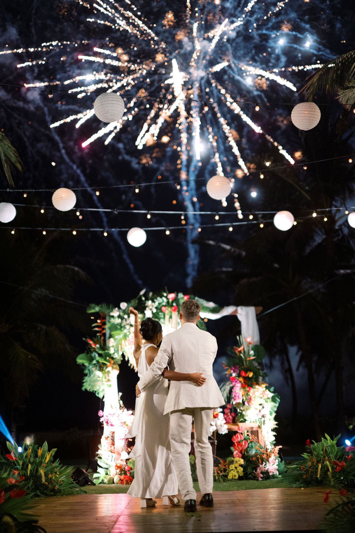Couple in white embracing under fireworks at tropical outdoor wedding with lanterns and floral backdrop at night.
