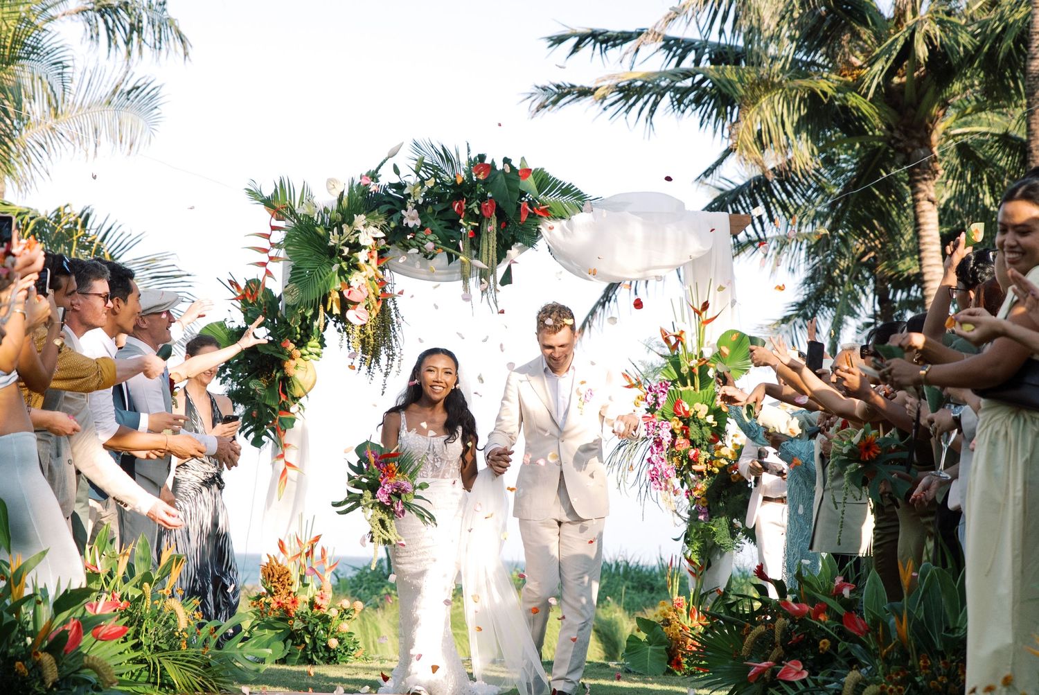 Two newlyweds walk under a tropical floral arch while guests celebrate with confetti at an outdoor wedding ceremony.