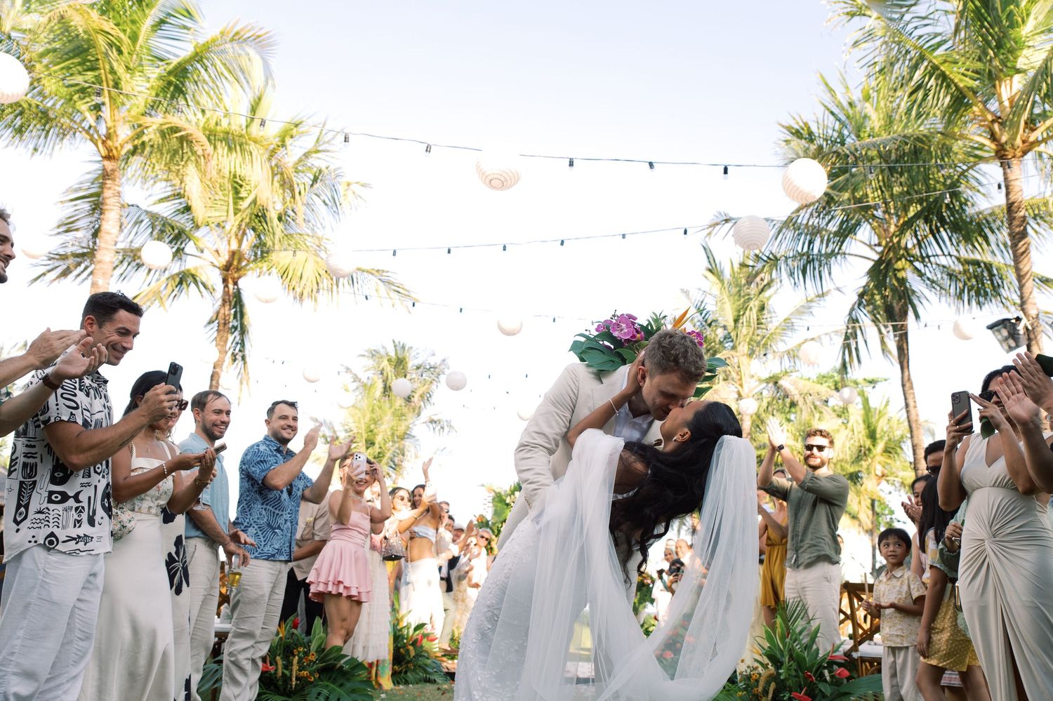 Couple shares a romantic kiss at tropical outdoor wedding ceremony with palm trees and cheering guests.