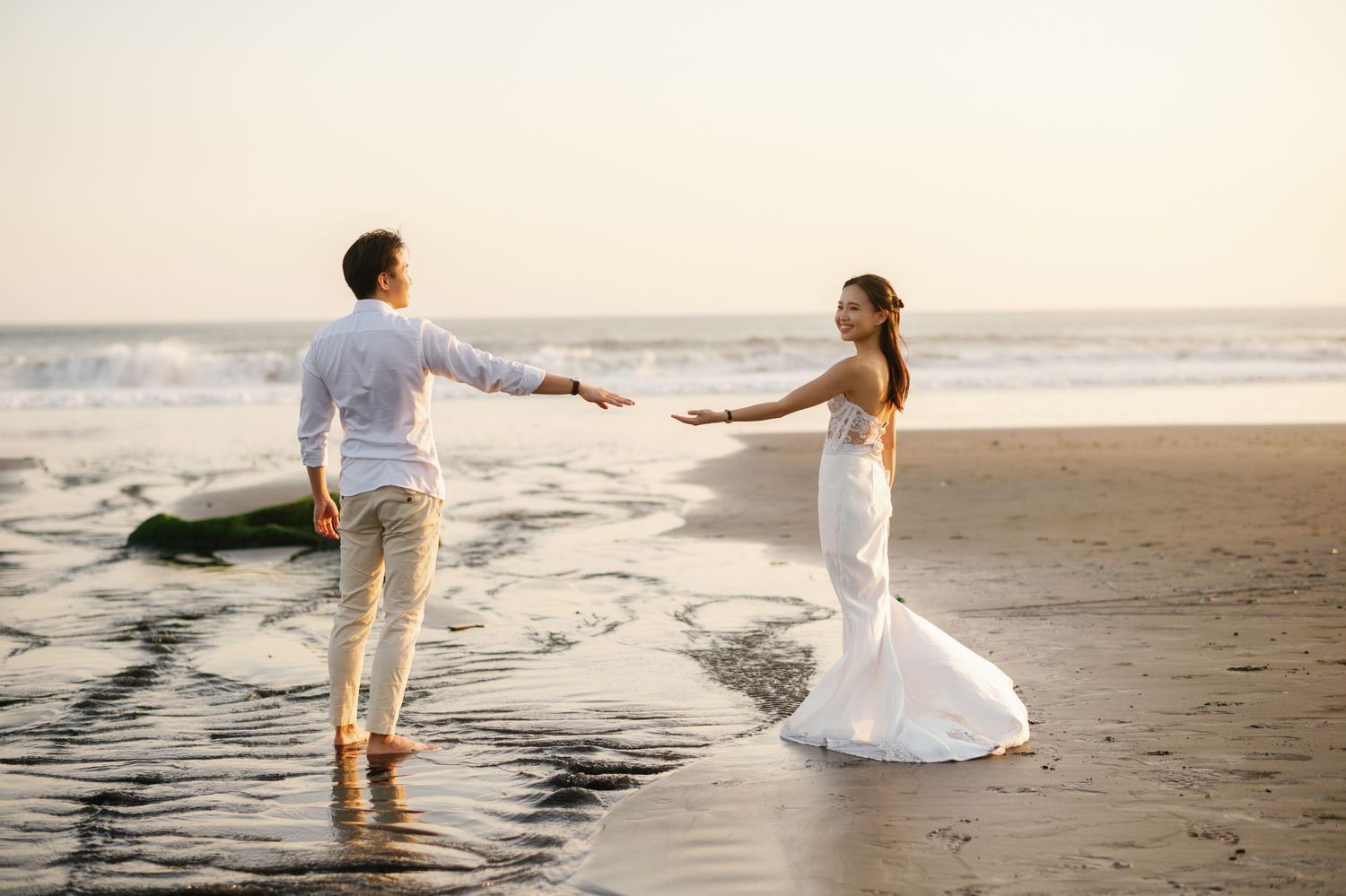Bride in white gown and groom in white shirt hold hands on a sunlit beach with ocean waves behind them.