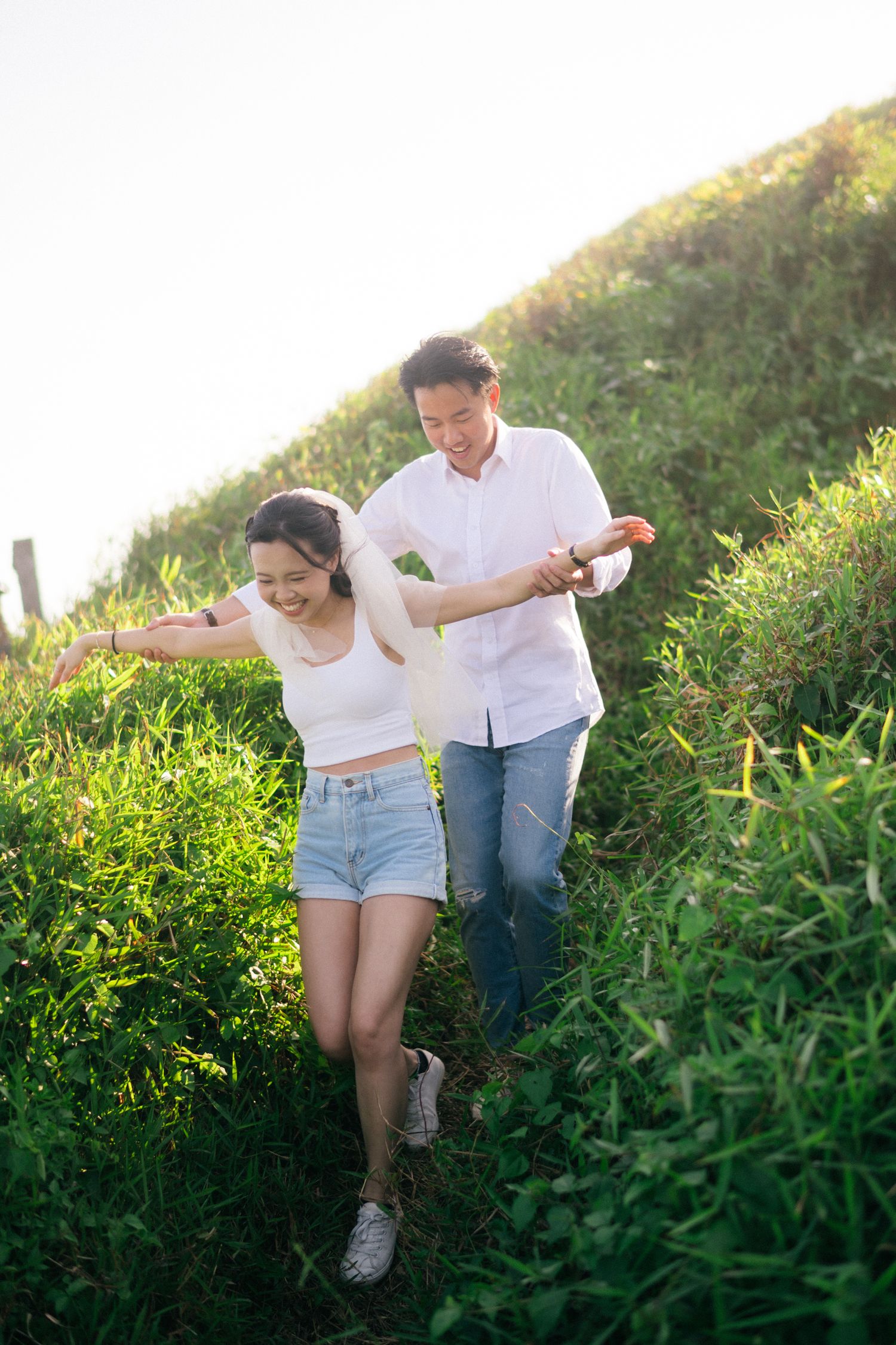 A joyful couple walks hand-in-hand through a lush green hillside path in warm sunlight.