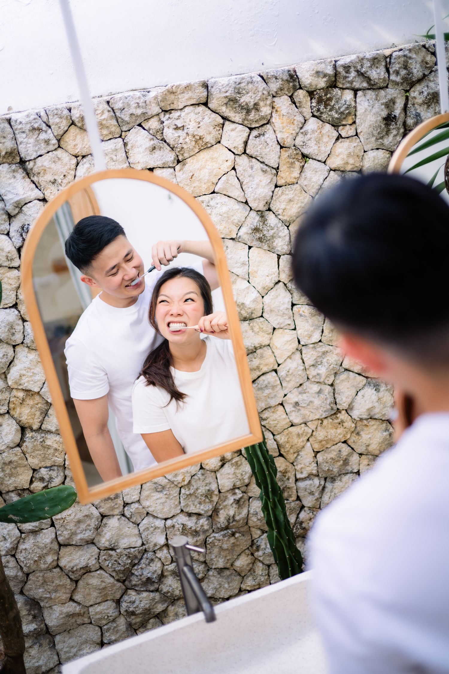 Asian couple brushing teeth together, smiling in arched mirror with stone wall background in modern bathroom.