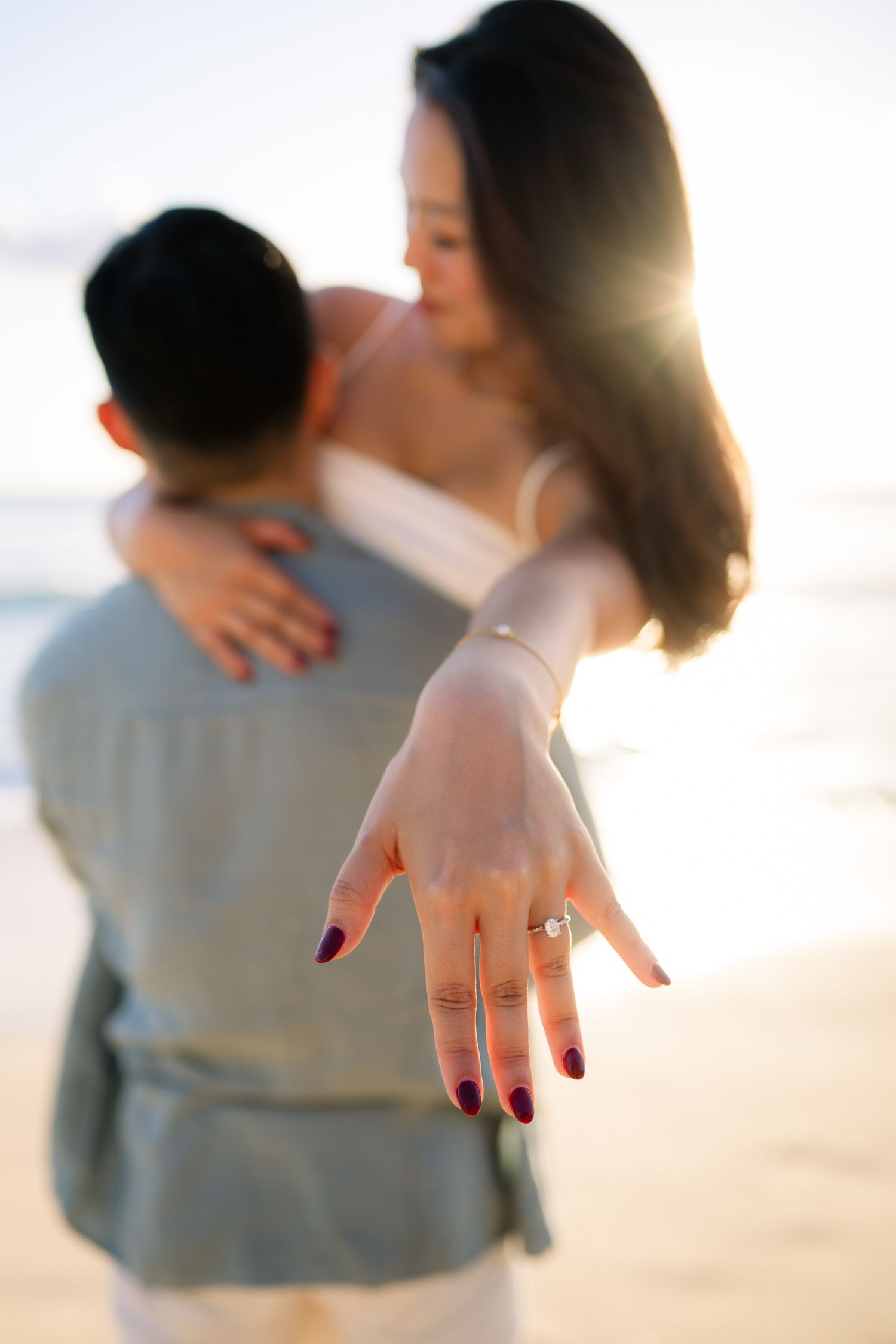 Couple embracing on beach at sunset, woman showing diamond engagement ring toward camera with dark red manicured nails.