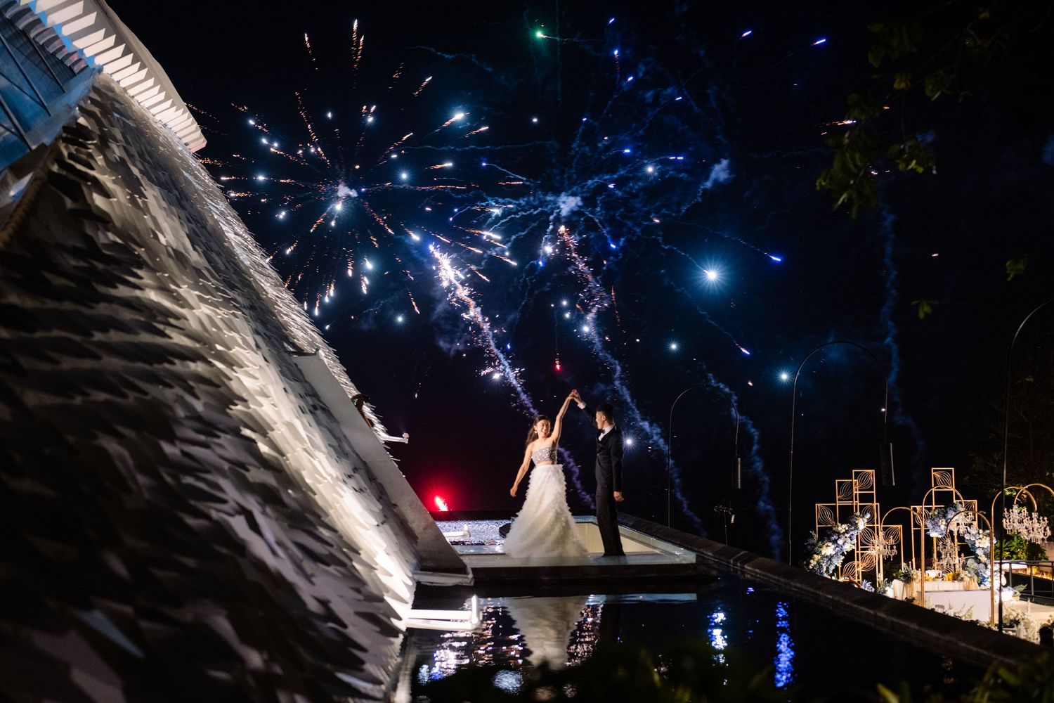 Bride and groom embrace on an illuminated wedding stage by the water as colorful fireworks burst across the night sky.