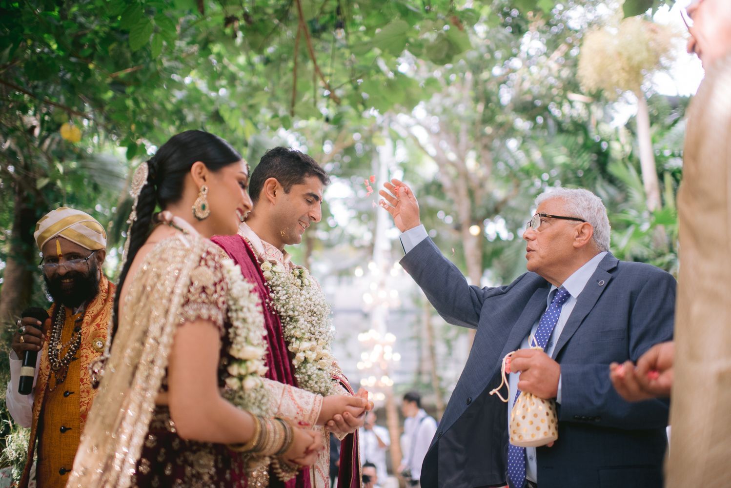 Elder man gestures during an Indian wedding ceremony as bride and groom stand adorned in traditional floral garlands outdoors.