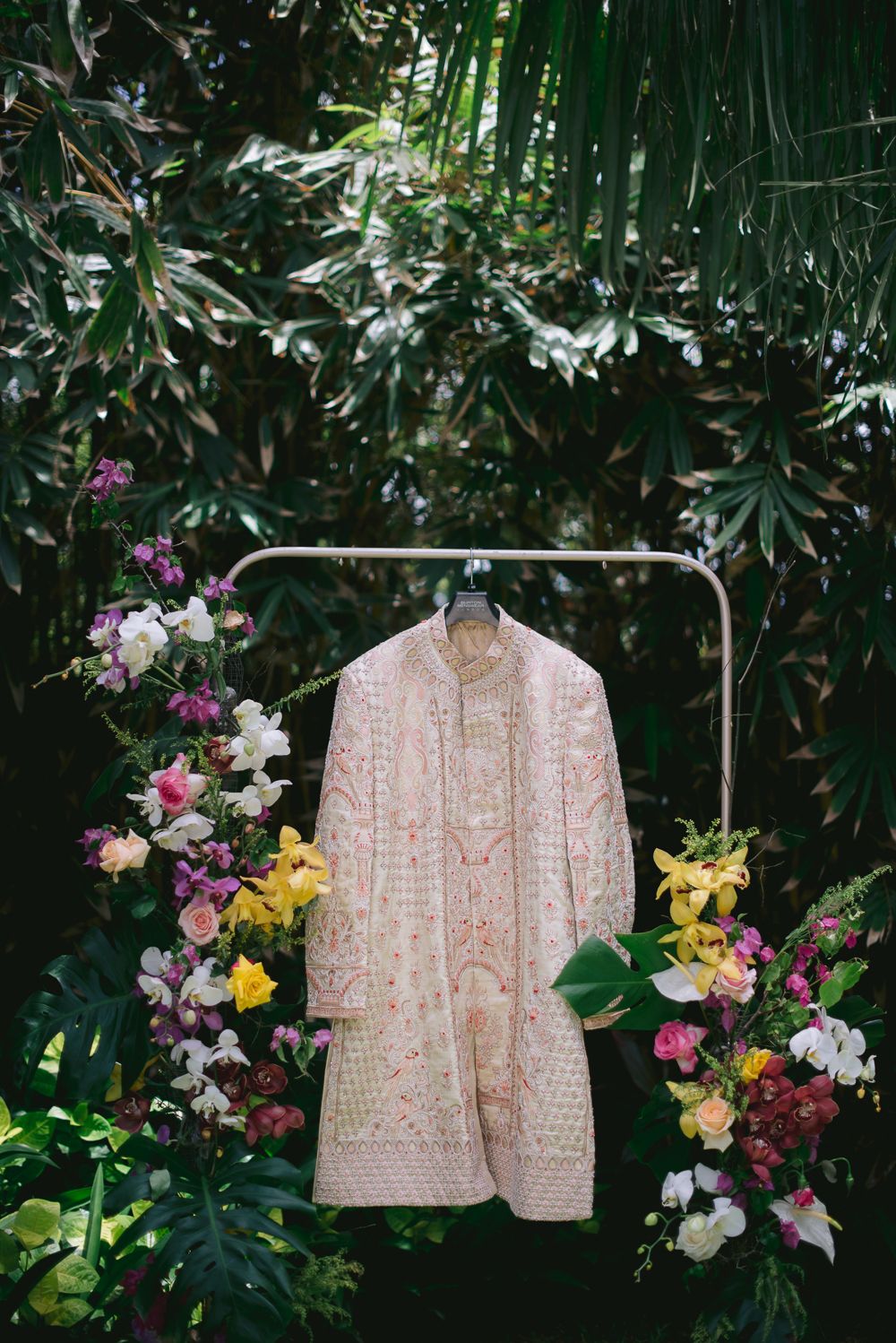 Pink embroidered sherwani displayed on a floral-adorned rack surrounded by lush greenery and colorful blooms.