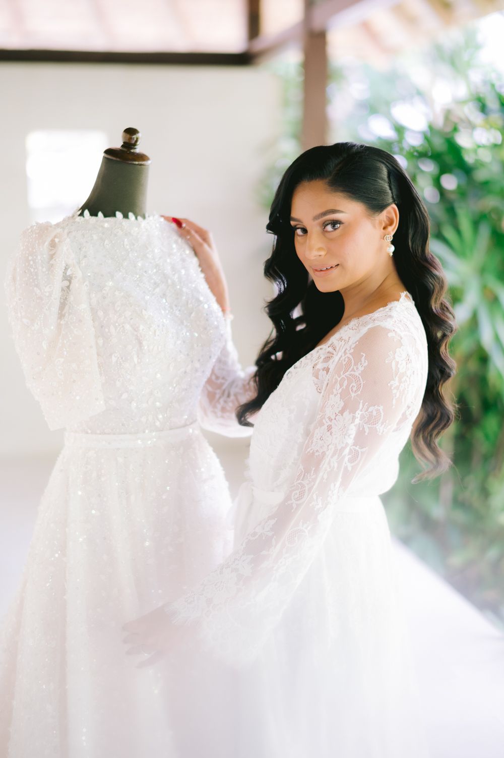 Bride in a beaded lace long-sleeve wedding gown adjusts dress on mannequin, smiling elegantly.