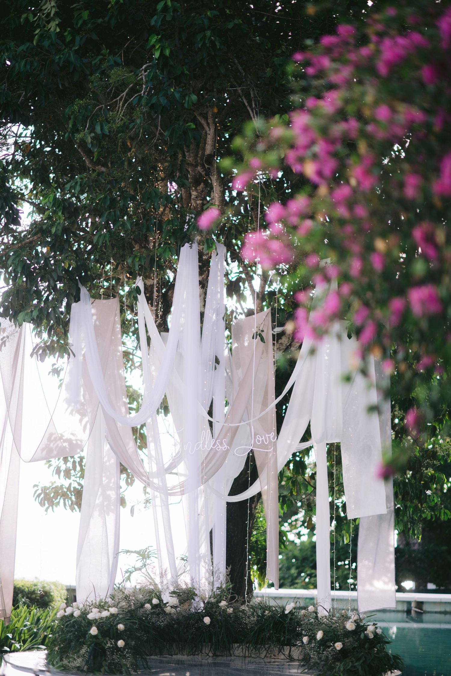 Elegant white fabric draping hanging from lush green trees at an outdoor wedding ceremony with pink flowers.