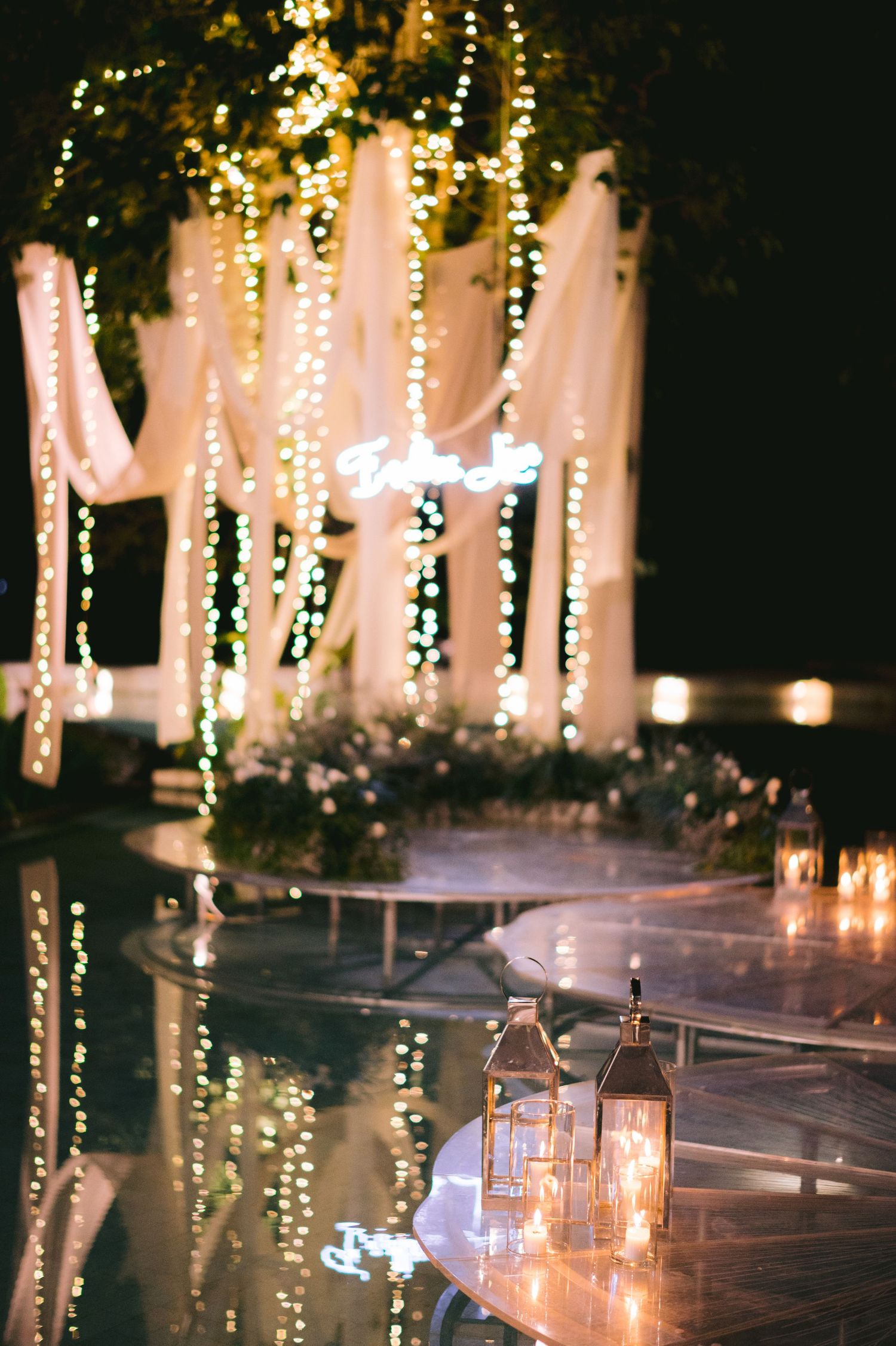Elegant outdoor wedding reception with fairy lights, white drapes, and lanterns reflecting on water at night.