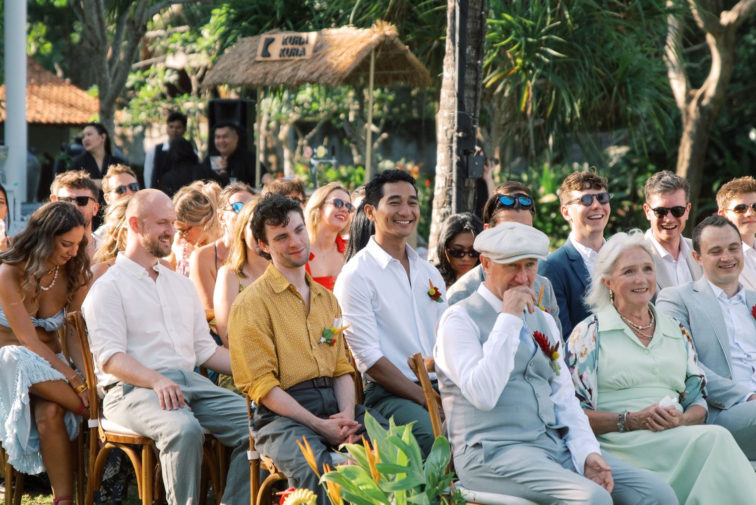Wedding guests smiling and laughing while seated outdoors at a tropical garden ceremony.