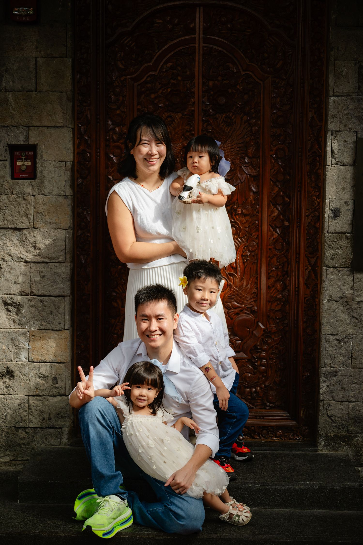 Happy Asian family of four posing in front of ornate wooden door, parents and two young children in white outfits.