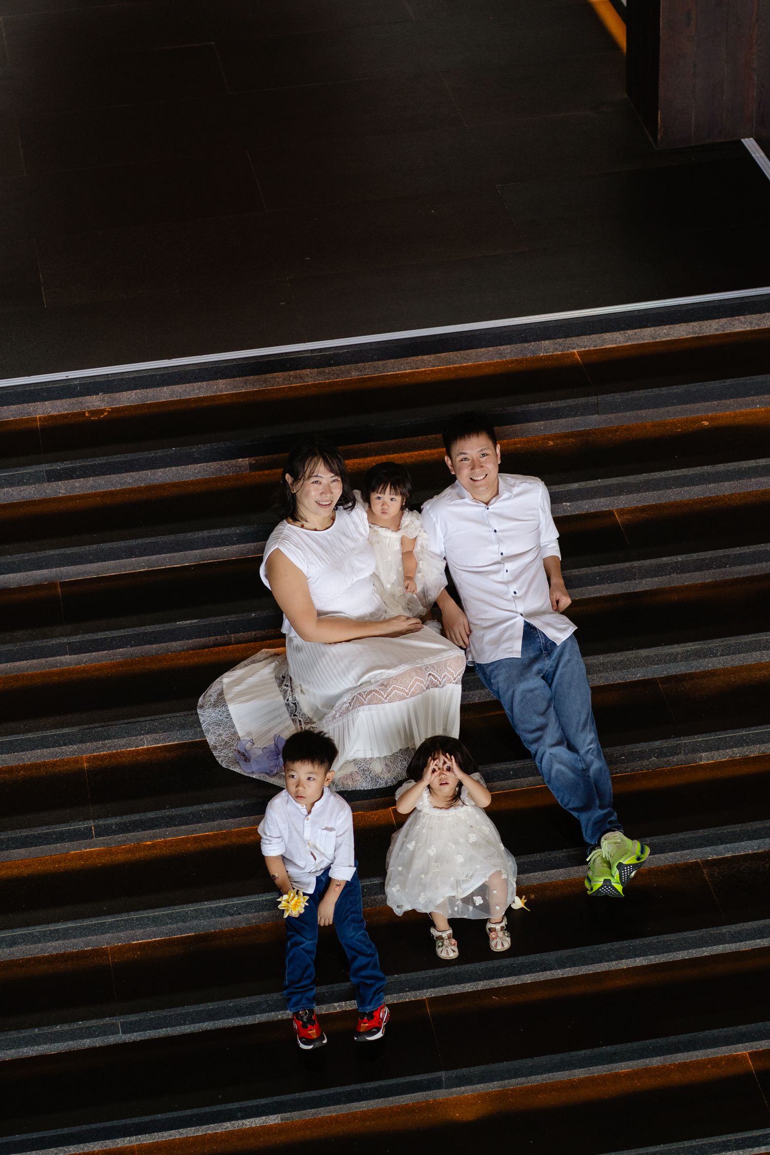 Family of five posing on wooden stairs, parents with three young children, dressed in white and blue outfits.