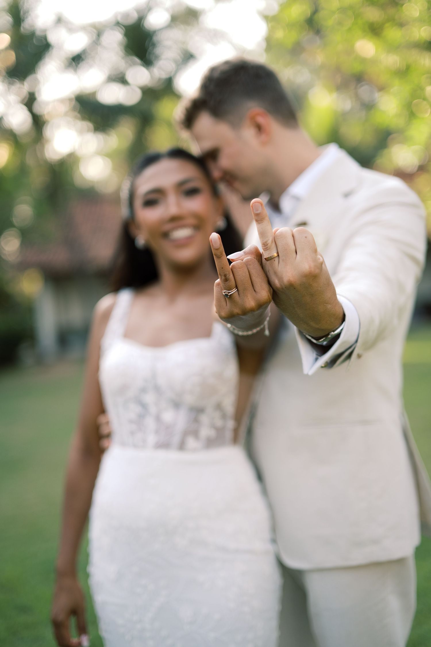 Newlywed couple showing off wedding rings, bride in white lace gown and groom in ivory suit, outdoors.