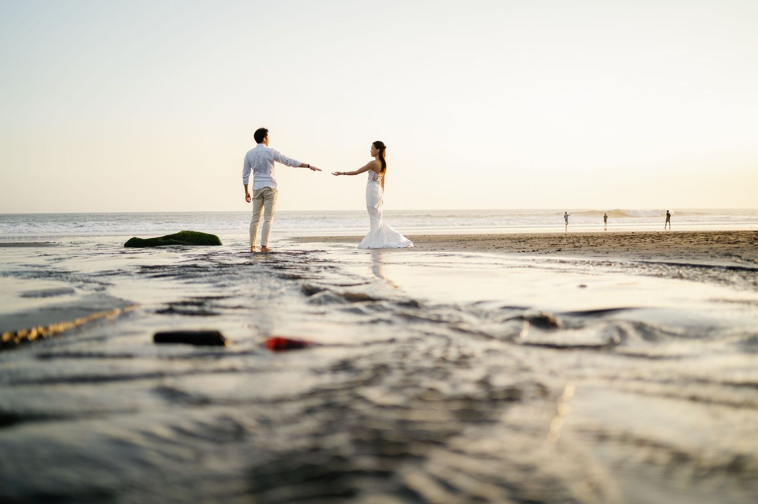 Couple holding hands on a wet beach at sunset in wedding attire.