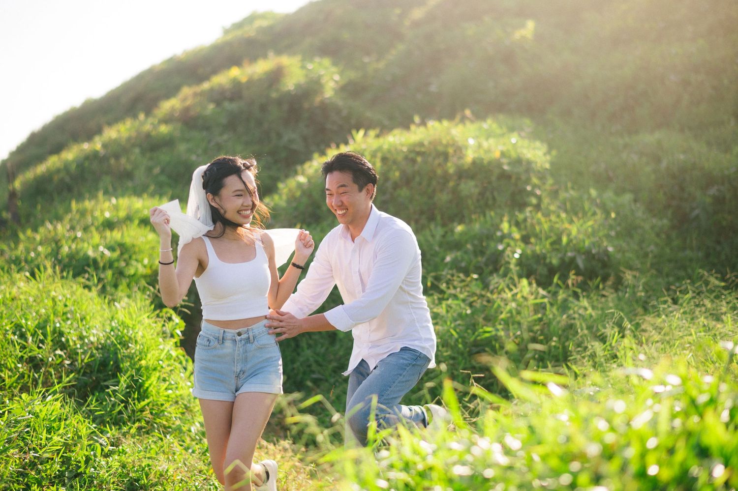 Couple holding hands laughing in sunny green meadow, woman wearing white crop top and denim shorts with veil.