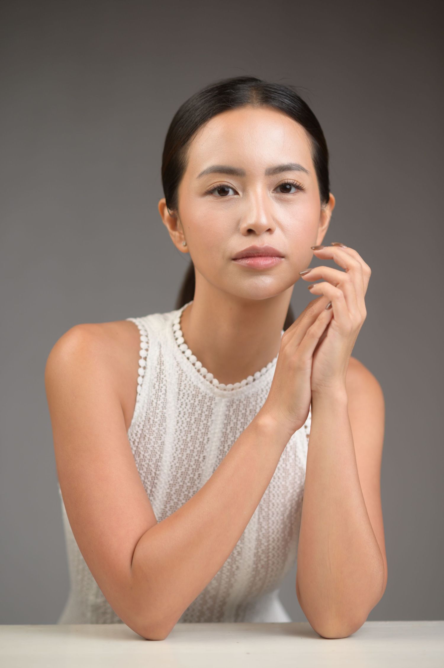 Young Asian woman in white lace sleeveless top poses gracefully with hands clasped near face against gray background.