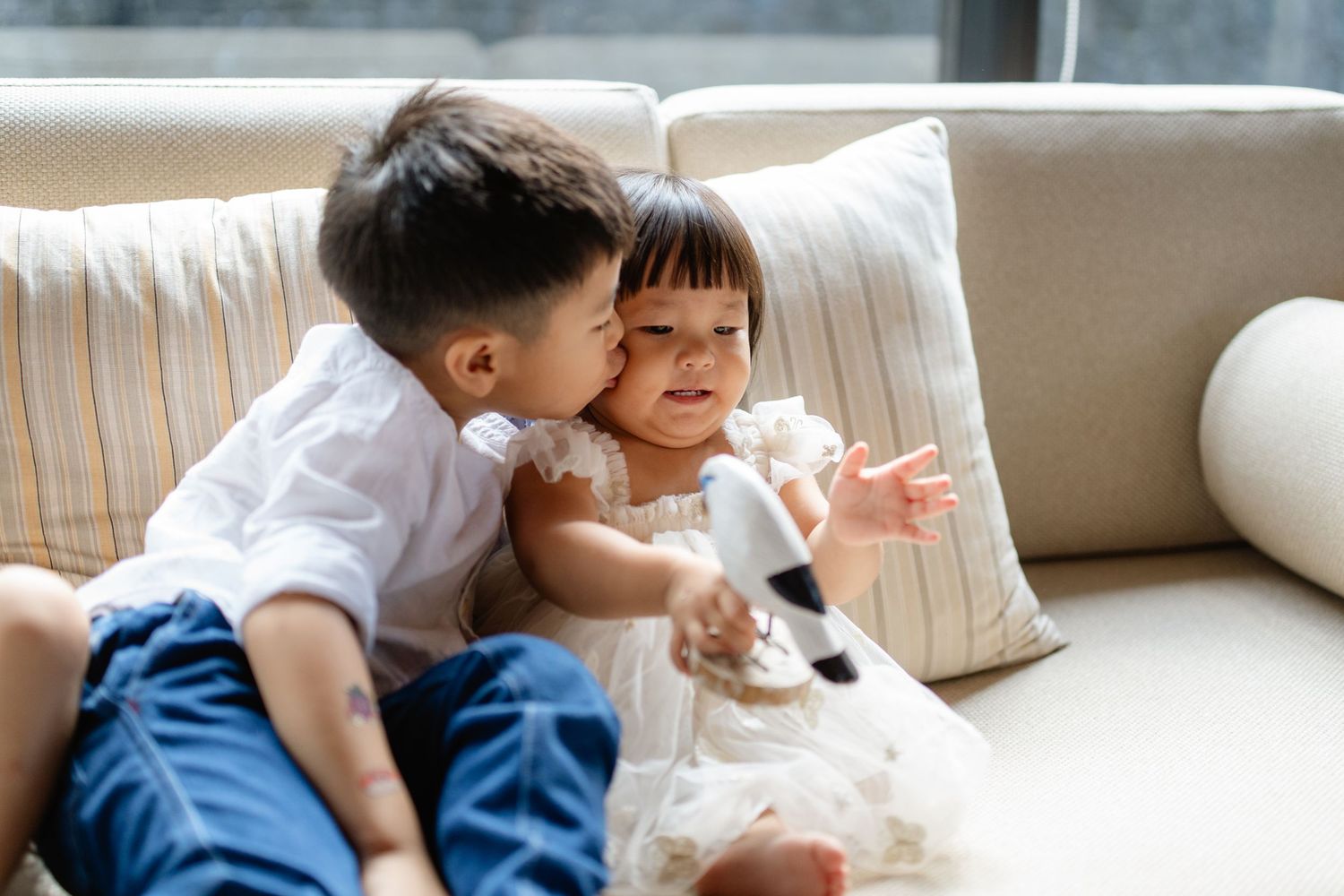 A young boy gently kisses his little sister on the cheek as they sit together on a beige sofa.
