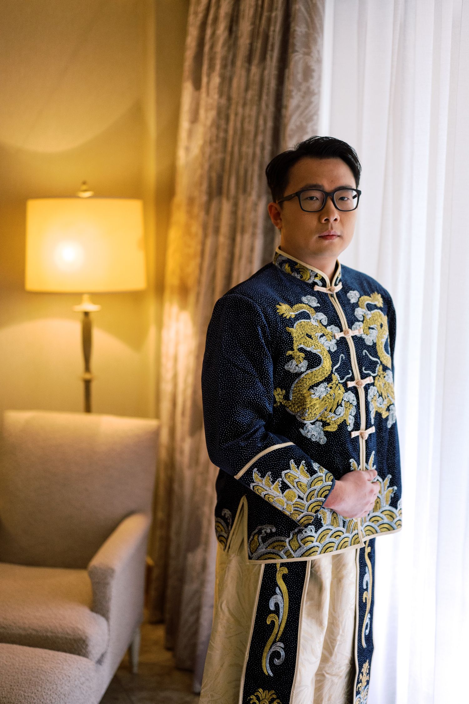 Man wearing traditional navy blue Chinese silk robe with golden dragon embroidery and glasses standing in elegant hotel room.