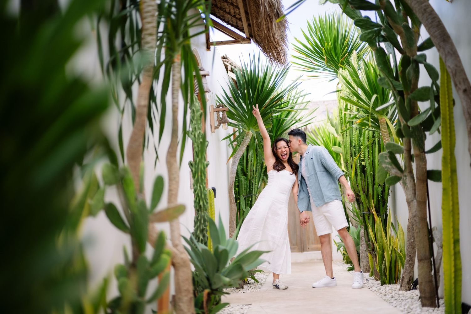 Couple in white outfits walking through a lush cactus garden pathway, sharing a romantic moment outdoors.