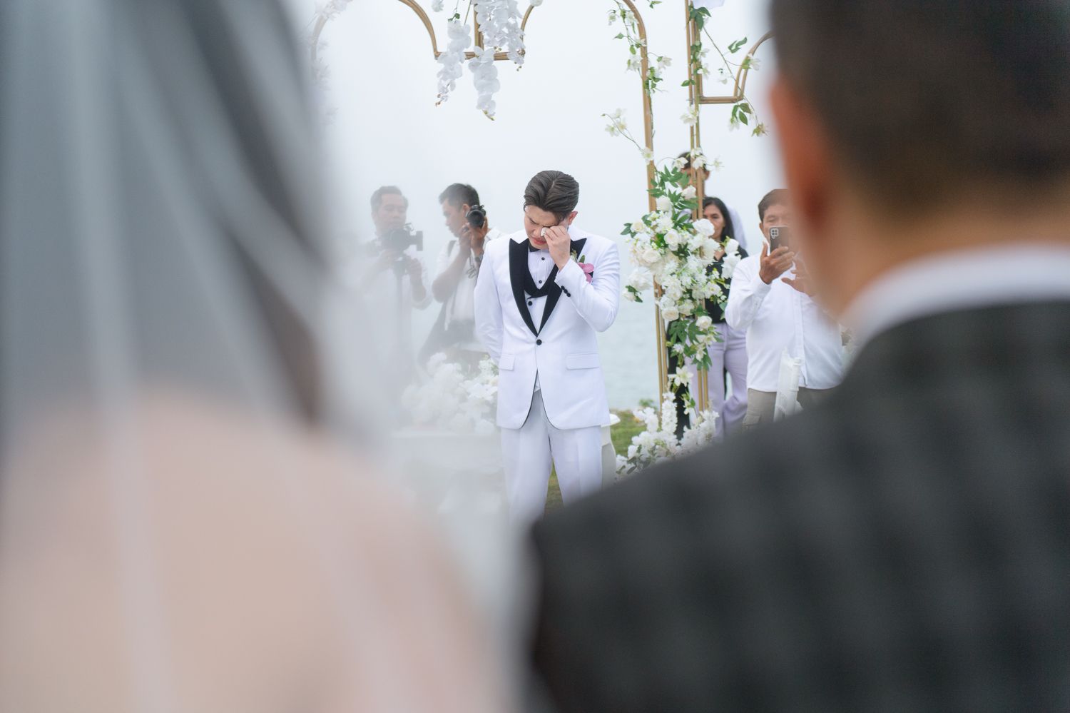 Groom in white tuxedo jacket wiping tears during an outdoor wedding ceremony with guests and floral arch.