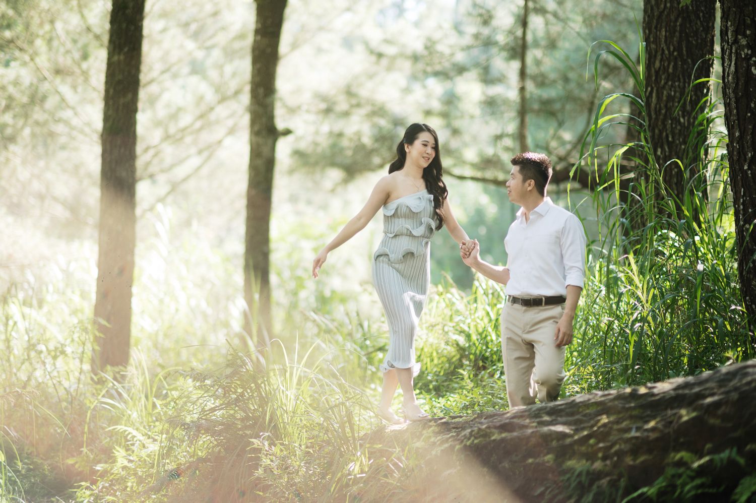 Couple holding hands walking through a sunlit forest with lush green trees and glowing natural light.