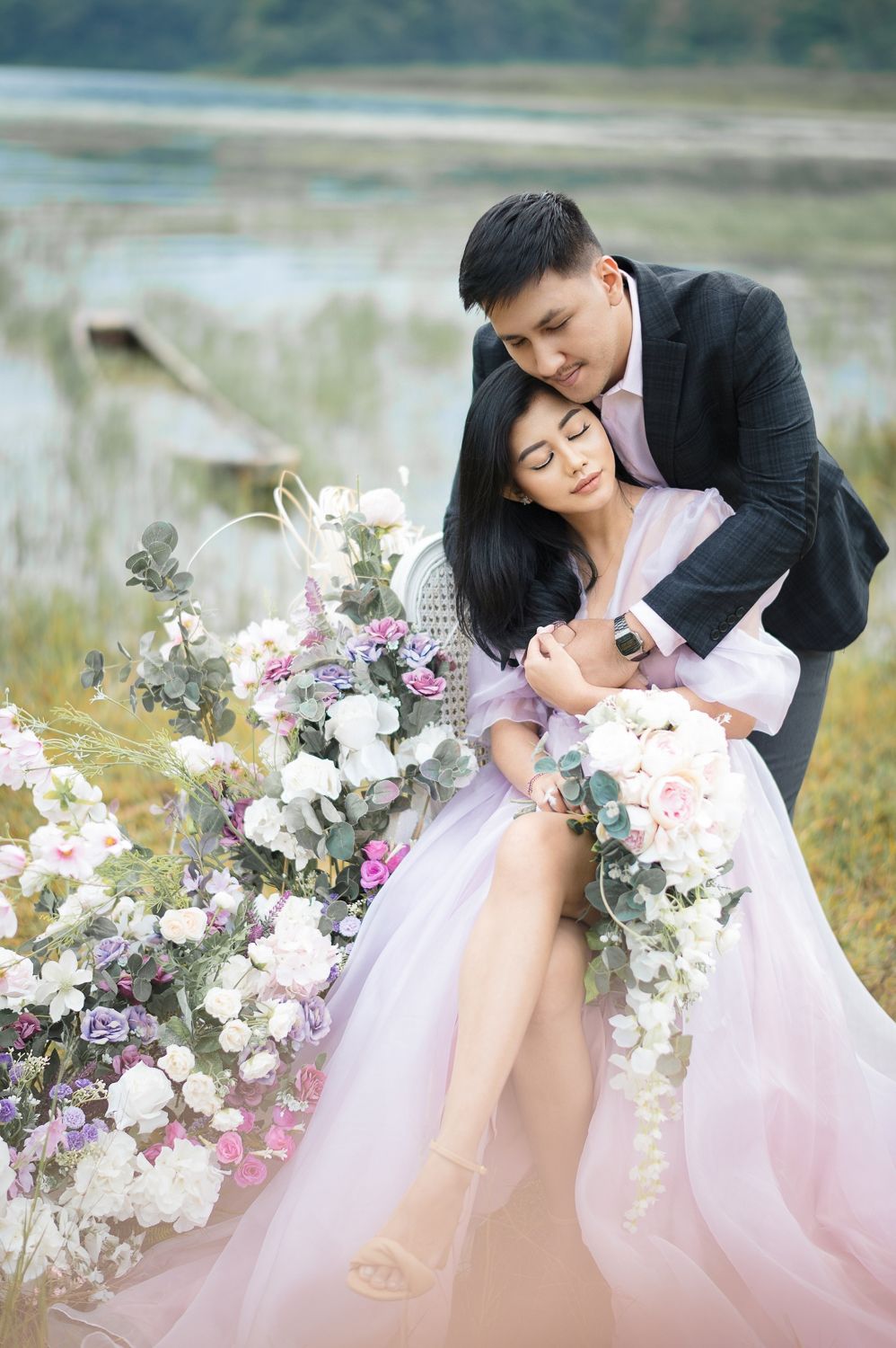 Couple in romantic embrace outdoors, woman in lavender gown holding bouquet, man in dark suit, surrounded by floral arrangements.