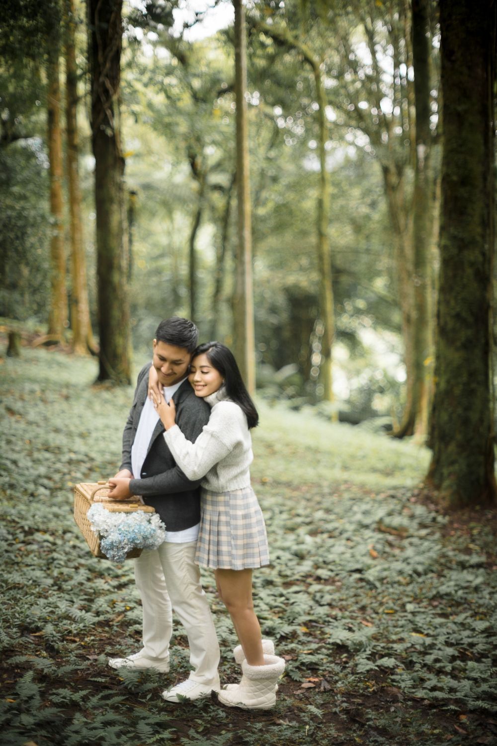Romantic couple embracing in a lush green forest, woman in plaid skirt holding blue flowers, man in gray blazer.