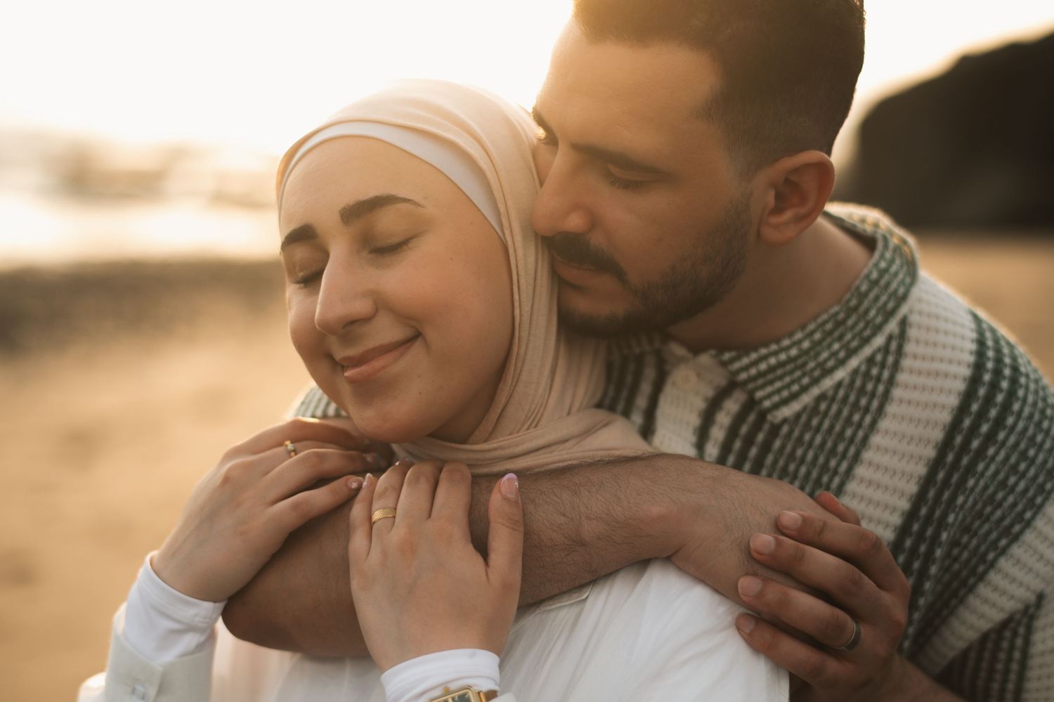 A man tenderly embraces a smiling woman wearing a hijab during a golden sunset outdoor moment.