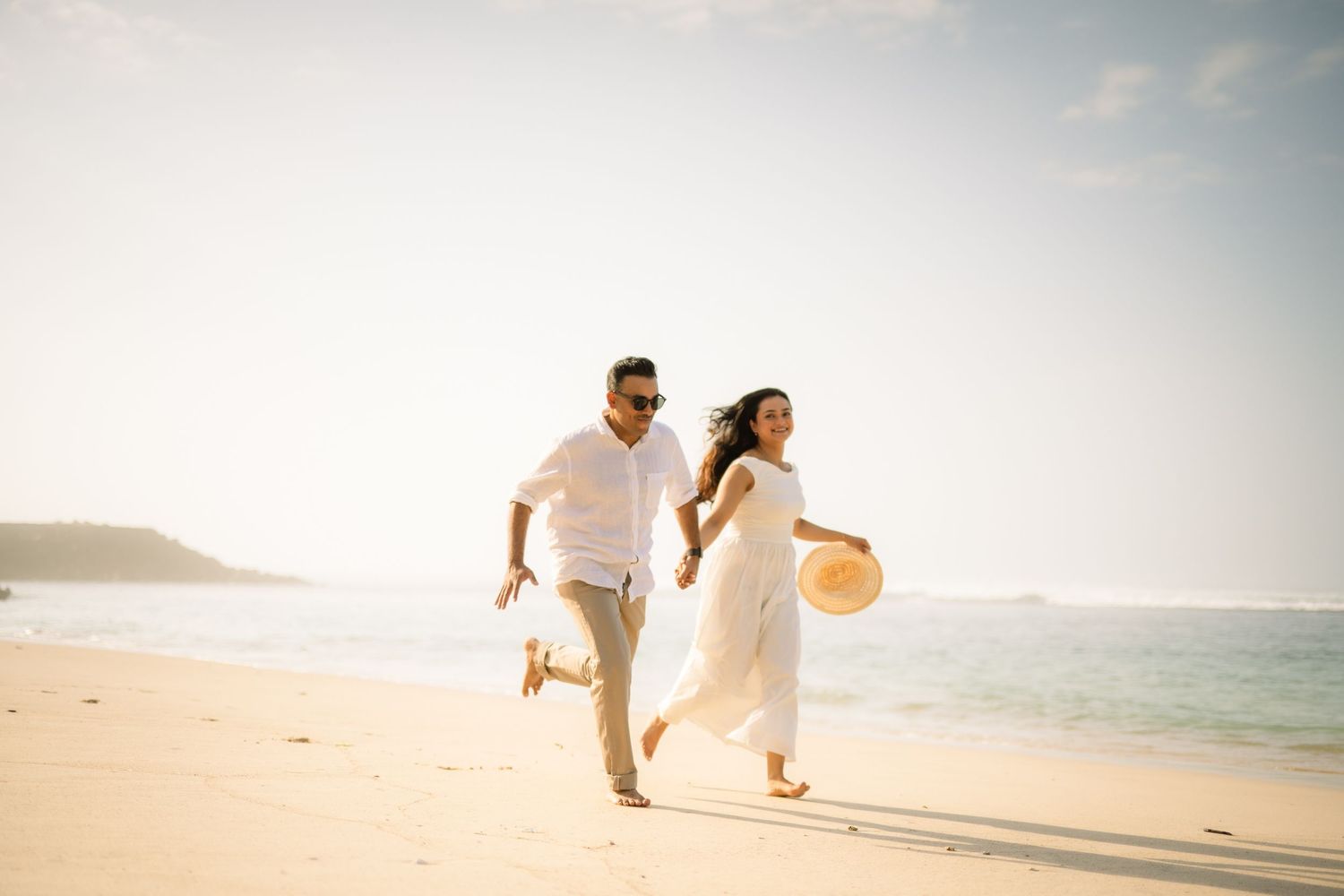 Happy couple in white outfits running on a sunny tropical beach, holding hands and smiling joyfully.