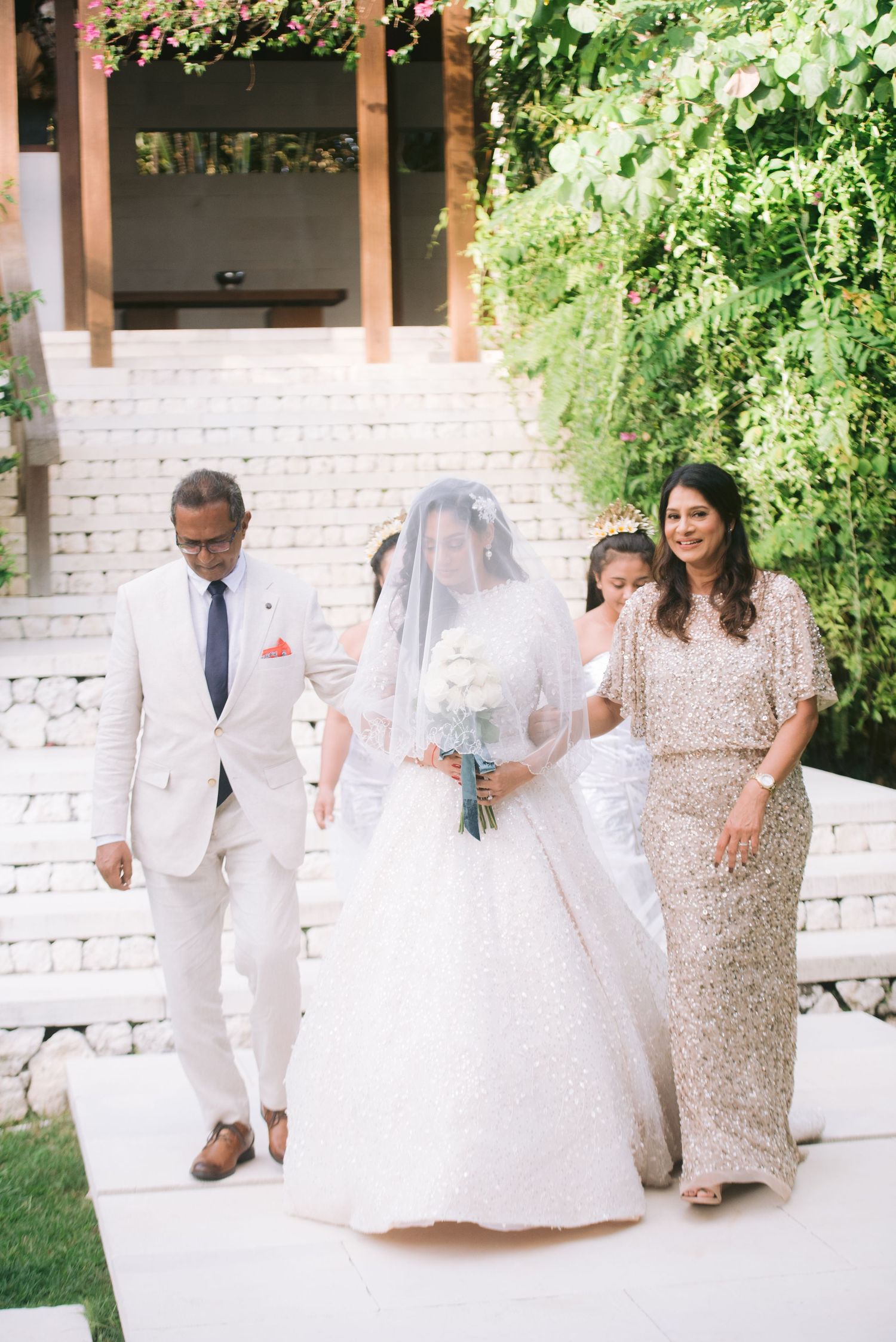 Bride in white gown and veil walks down the aisle with parents at an outdoor garden wedding ceremony.