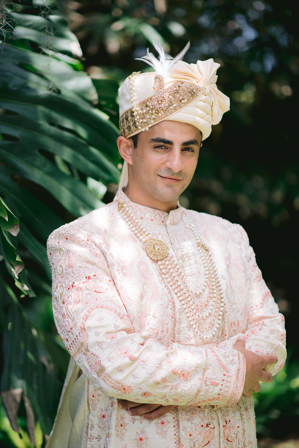 Groom in traditional Indian sherwani with gold embroidery, pearl necklaces, and ornate turban with feather, arms crossed.