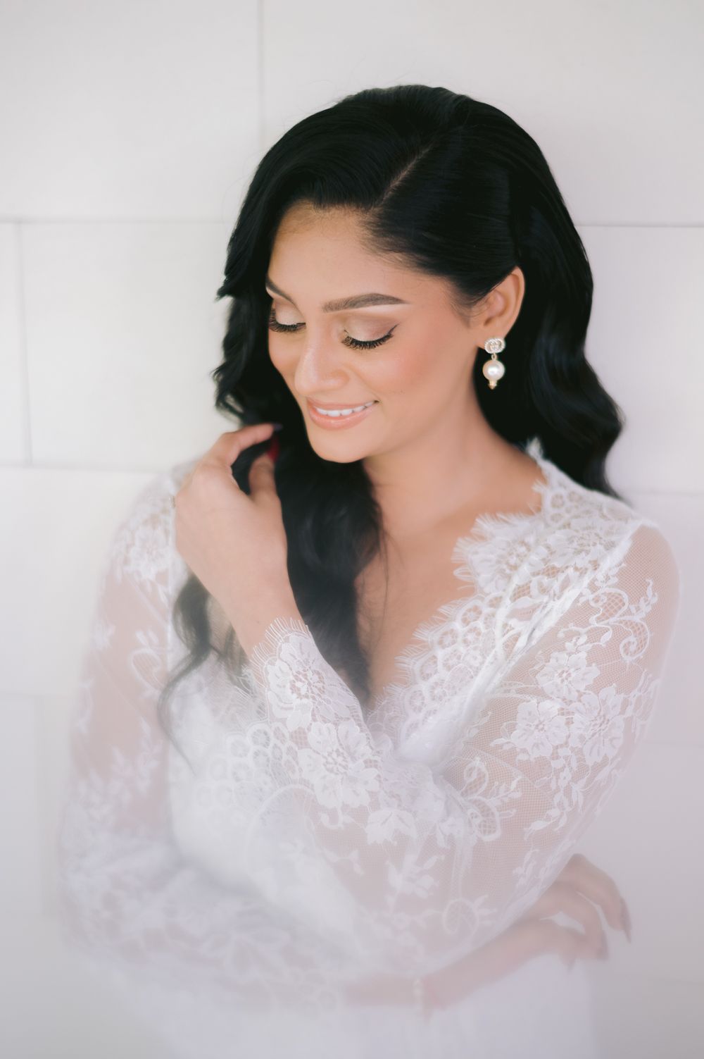 Elegant bride in white lace robe with pearl earrings, dark wavy hair, soft bridal makeup, posing gracefully indoors.