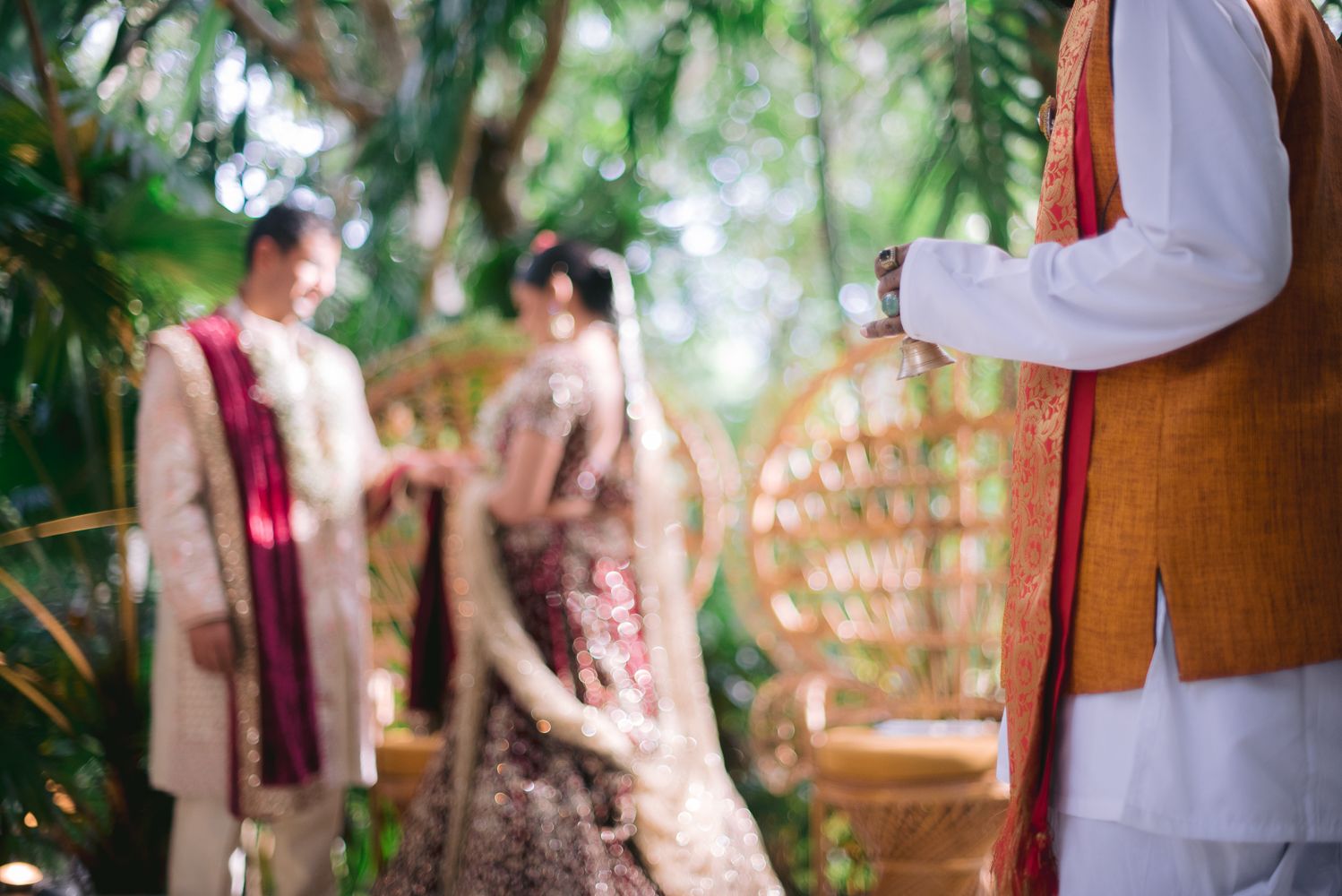 Indian wedding couple in traditional attire standing outdoors amid lush greenery with a decorative peacock chair.