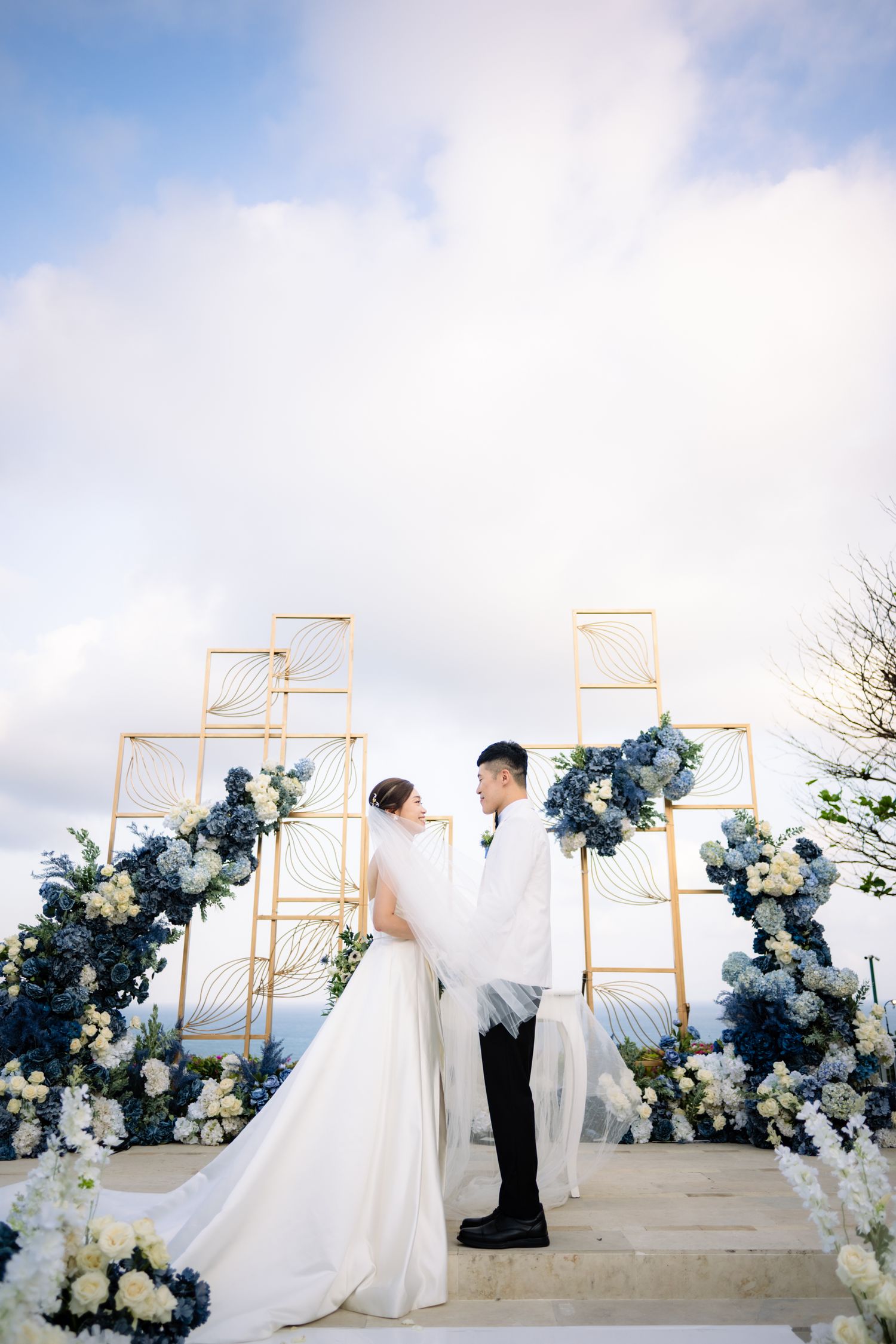 Bride and groom stand face to face under a veil at a cliffside wedding ceremony with blue and white floral arrangements and gold geometric a