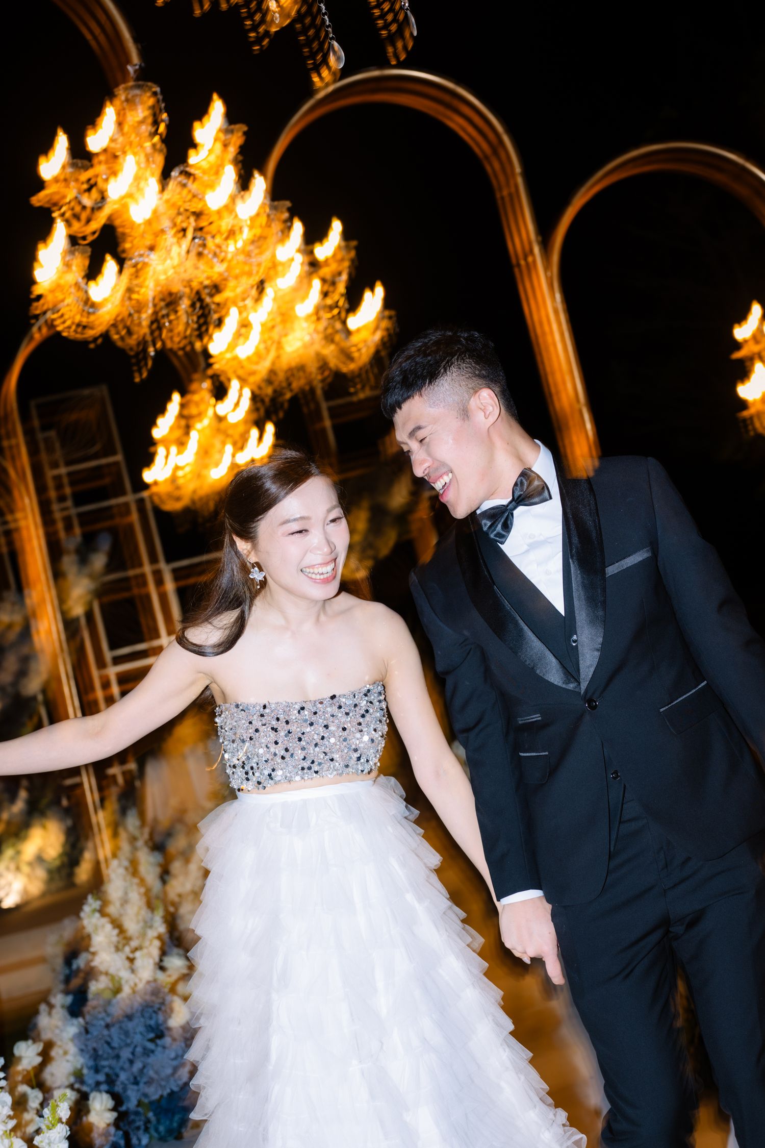 Smiling bride in a sparkling strapless tulle gown laughs with groom in a black tuxedo beneath glowing chandeliers at an evening wedding.