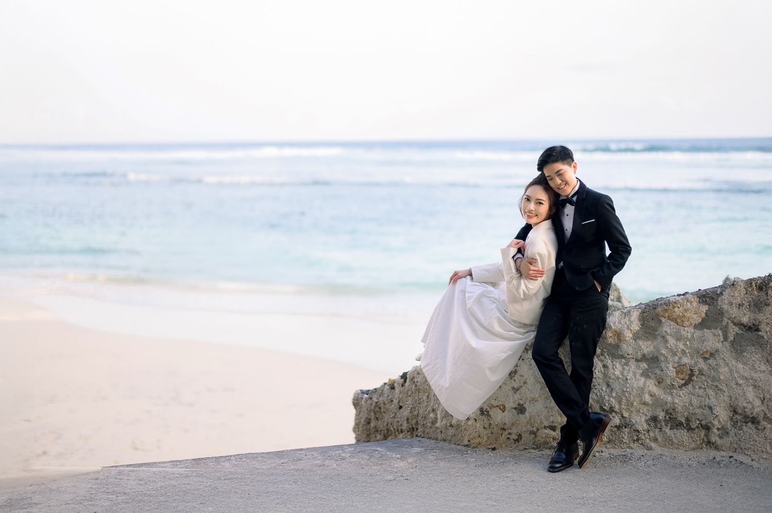 Couple in wedding attire posing on rocky beach wall with ocean backdrop, groom in black suit, bride in white gown.